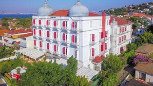 Un edificio blanco con contraventanas rojas y dos torres abovedadas, rodeado de vegetación y otros edificios.