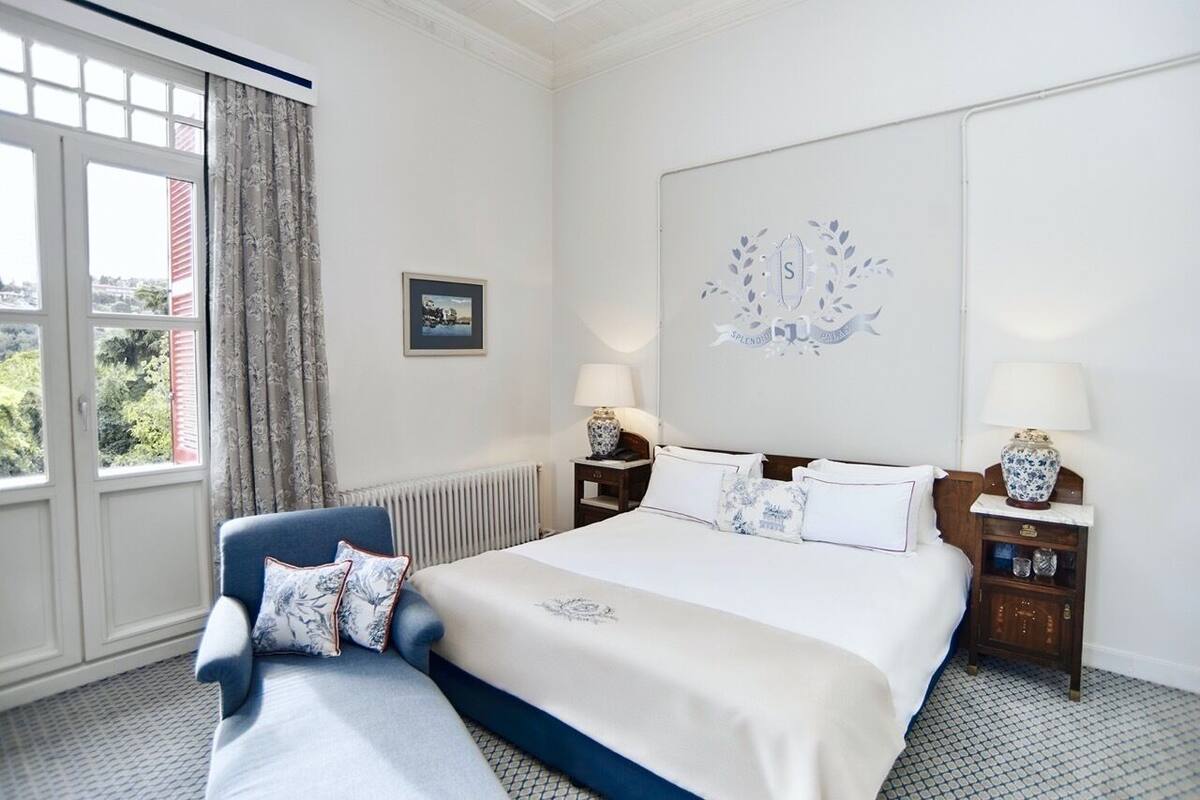 Une chambre d’hôtel avec un grand lit, un fauteuil bleu, deux tables de chevet avec des lampes, une grande fenêtre avec des rideaux et un tableau encadré au mur.