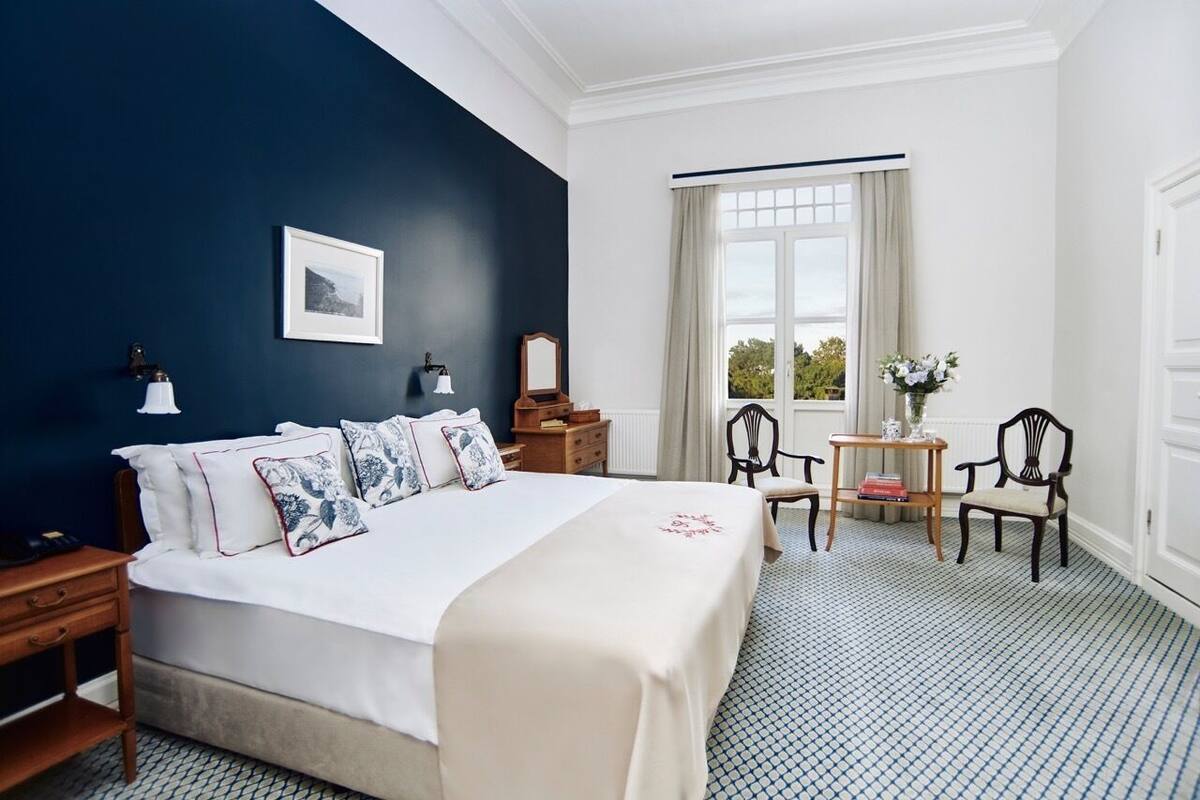 Une chambre à coucher avec un grand lit, des tables de chevet, une commode, une fenêtre avec des rideaux et une petite table sur laquelle est posé un vase.