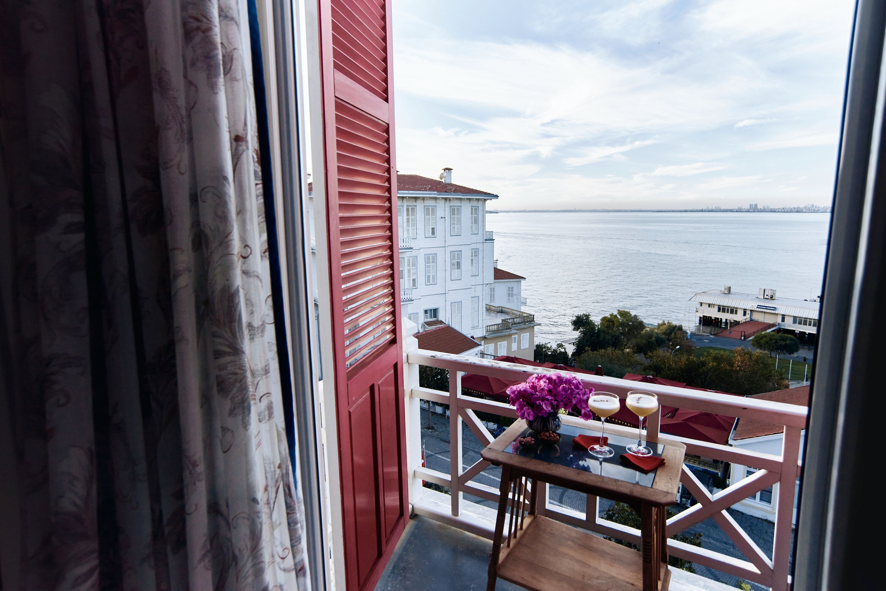 A balcony with a view of the sea, a table with drinks, and a vase of flowers.