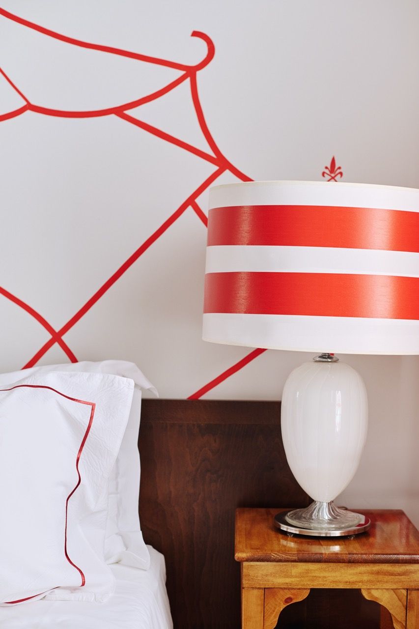 A neatly made bed with a white comforter and red piping, a wooden nightstand with a white lamp featuring red stripes, and a wall with a geometric red design.