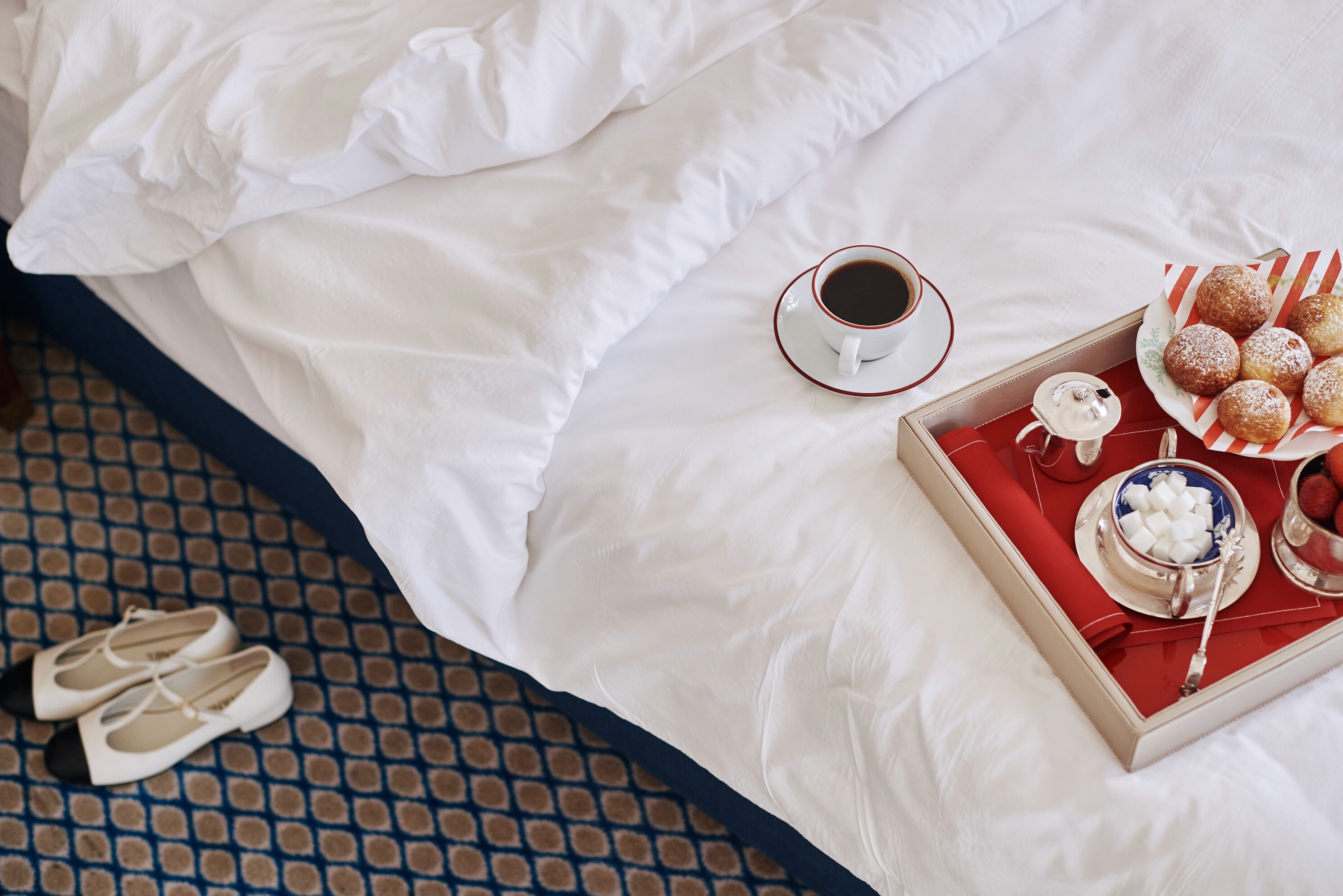 A neatly made bed with a pair of shoes, a cup of coffee, and a tray with pastries.