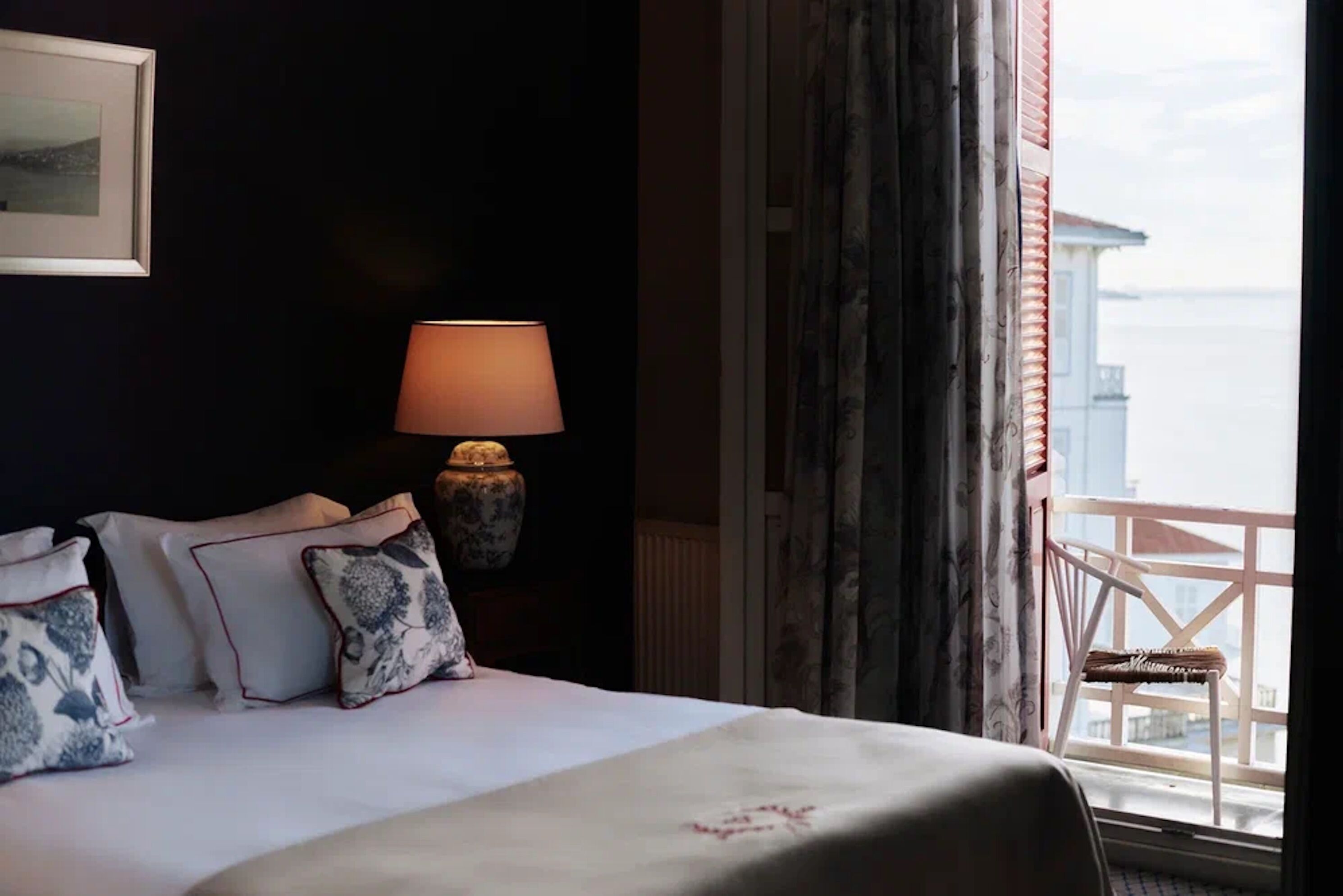 A hotel room with a bed, a bedside table with a lamp, a window with a view of buildings, and a balcony with a chair.