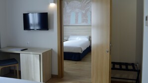 A bedroom with a bed, a desk with a chair, a TV mounted on the wall, and a door leading to another room. - Hotel Balneario de Graena (Cortes y Graena)