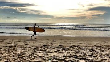 Una playa con huellas que conducen al agua, un surfista cargando una tabla de surf, y el océano bajo un cielo nublado.