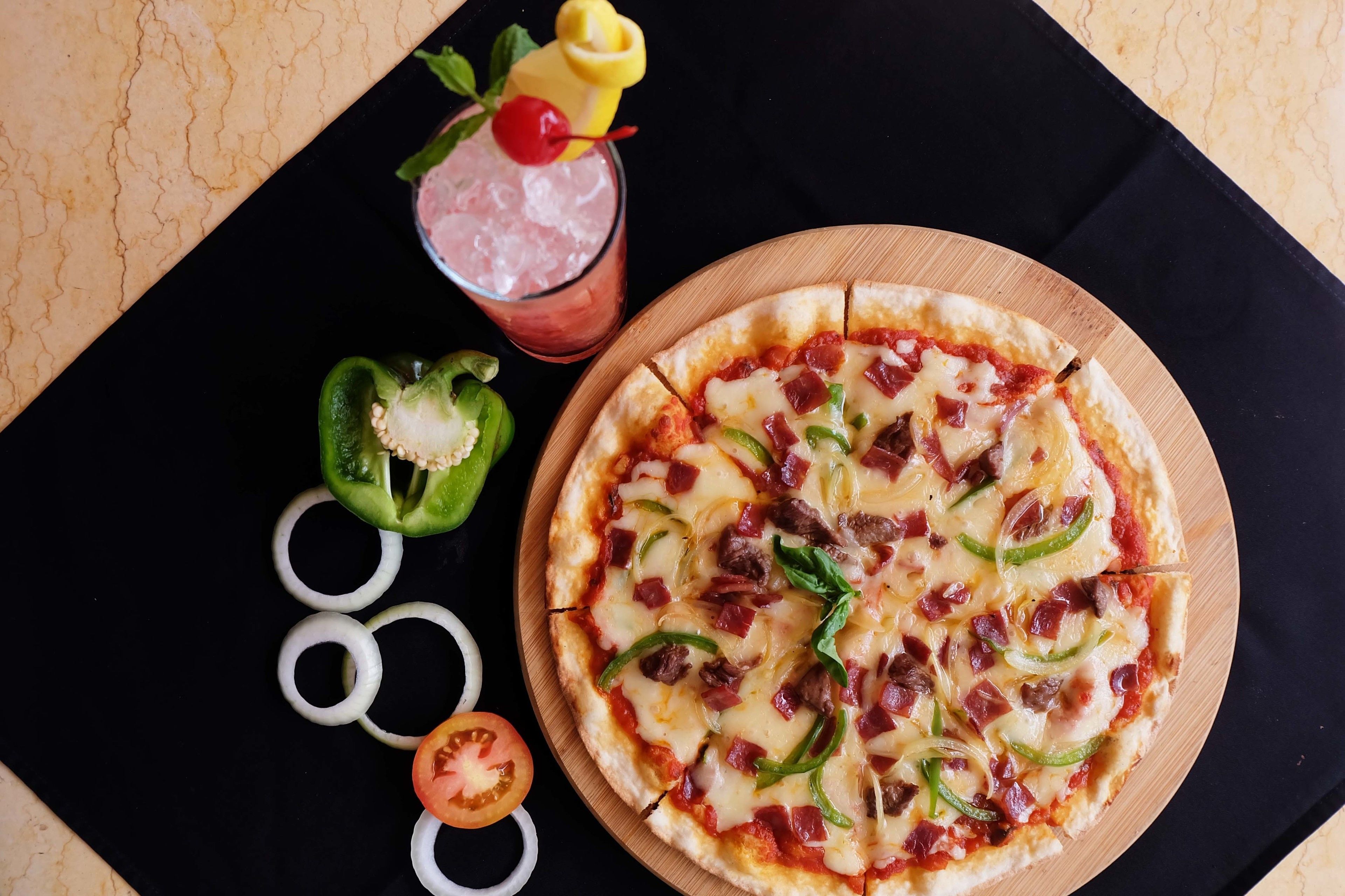 A pizza with various toppings, a drink with a cherry, and sliced vegetables on a wooden board.