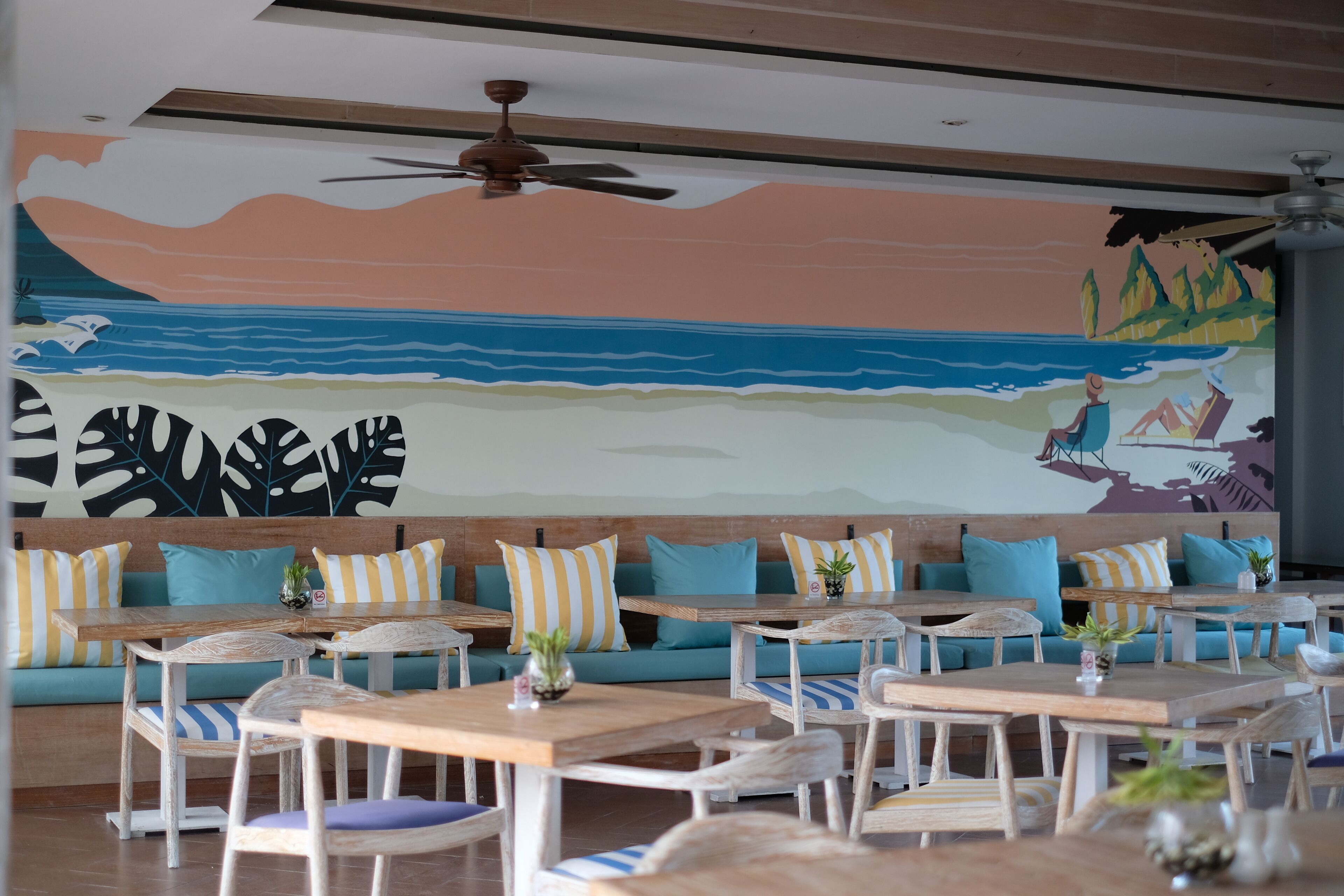 A restaurant with wooden tables and chairs, teal sofas with striped cushions, and a mural of a beach scene on the wall.