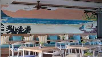 A restaurant with wooden tables and chairs, teal sofas with striped cushions, and a mural of a beach scene on the wall.