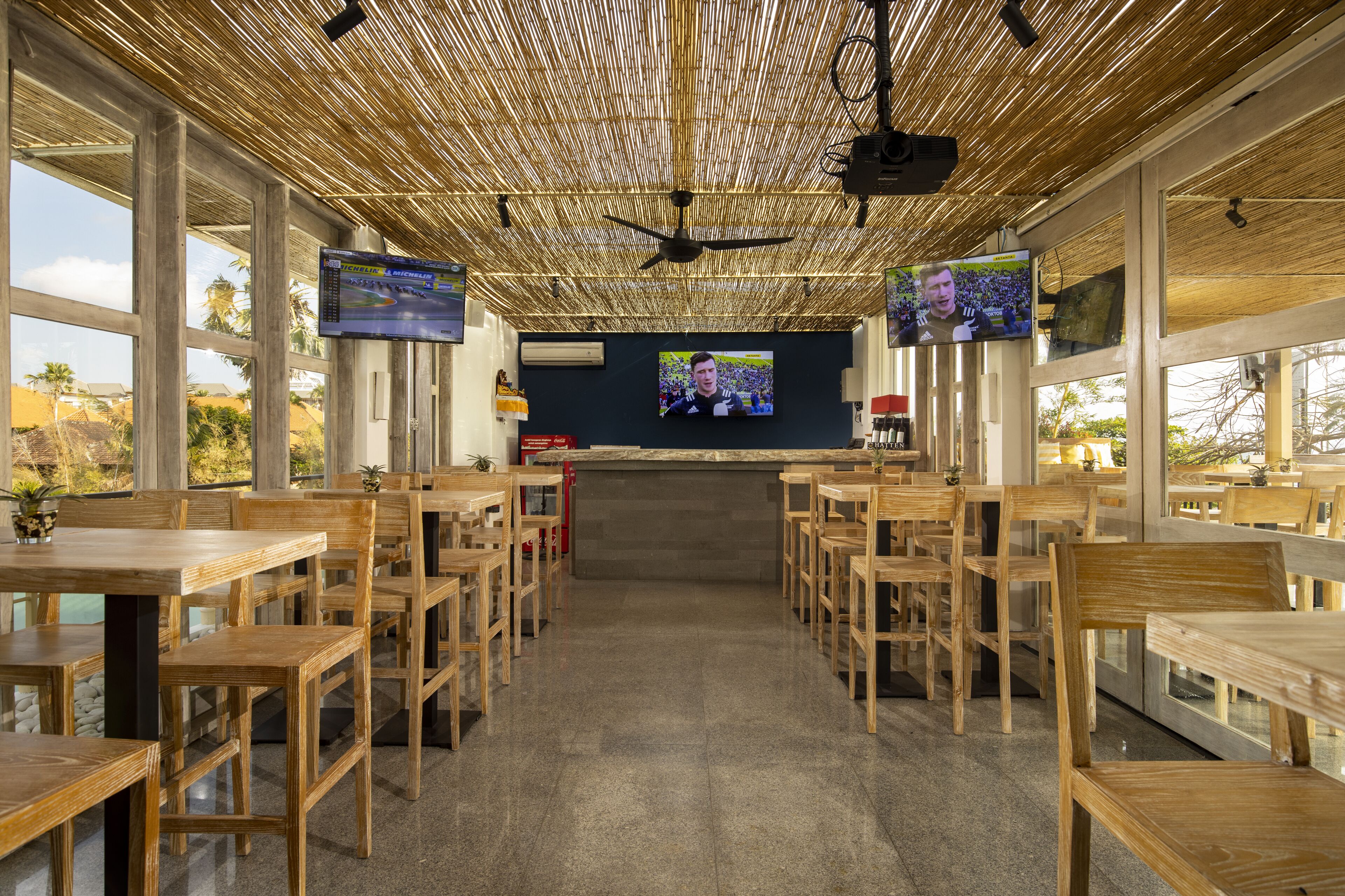 A restaurant with wooden tables and chairs, large windows, and a bamboo ceiling.