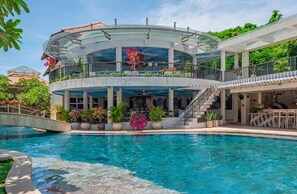 Un edificio moderno con piscina, que destaca por un nivel superior con paredes de vidrio, zona de estar al aire libre y un área cubierta con escalera.