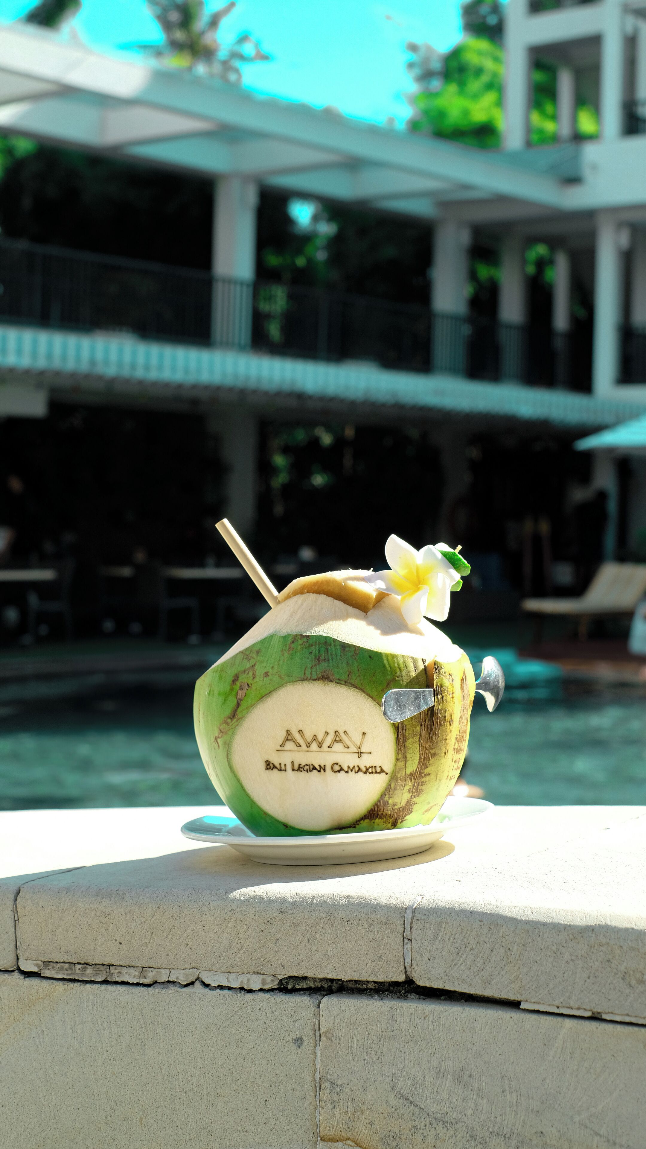 A coconut drink with a straw and a flower on a table near a pool.