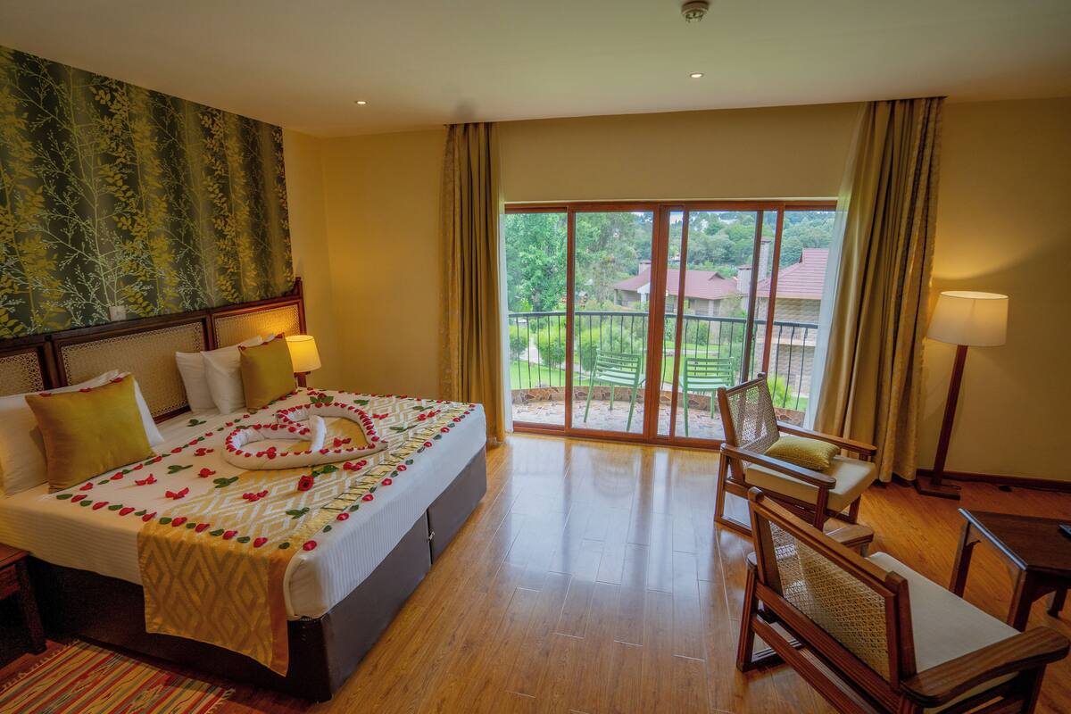 A hotel room with a bed, a chair, a table, and a view of greenery outside.