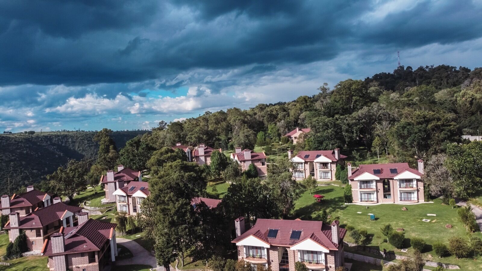 A residential area with houses featuring red roofs and brick exteriors, surrounded by lush greenery and hills.