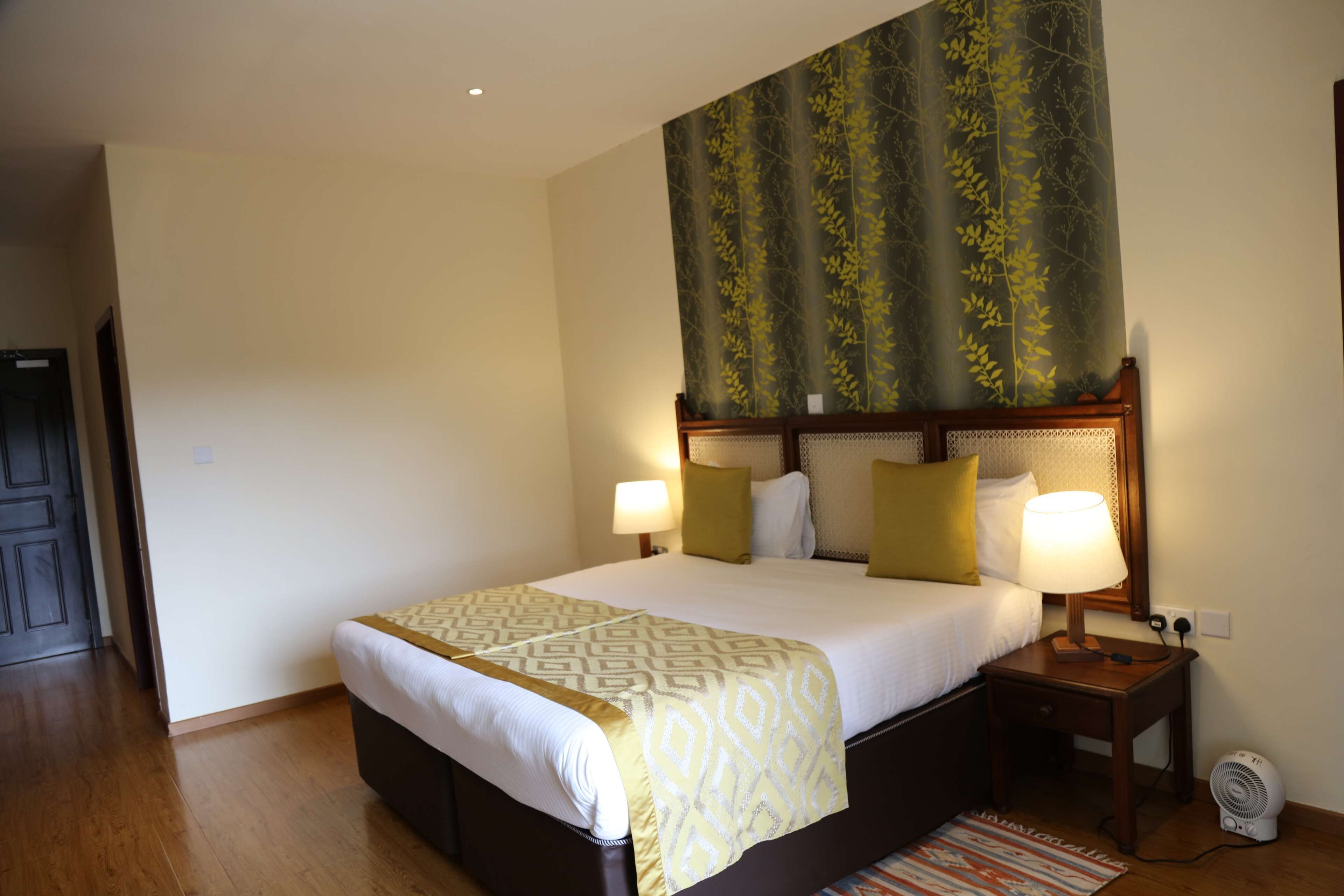 A hotel room with a large bed, two bedside lamps, a nightstand with a lamp, a wall-mounted fan, and a patterned tapestry above the bed.