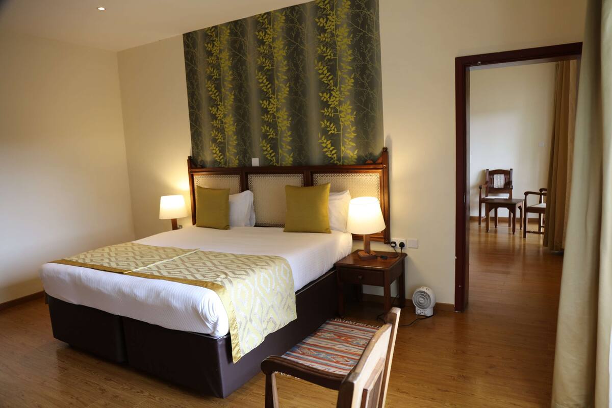 A hotel room with a large bed, two bedside tables with lamps, a wooden bench, and a view of another room through an open door.
