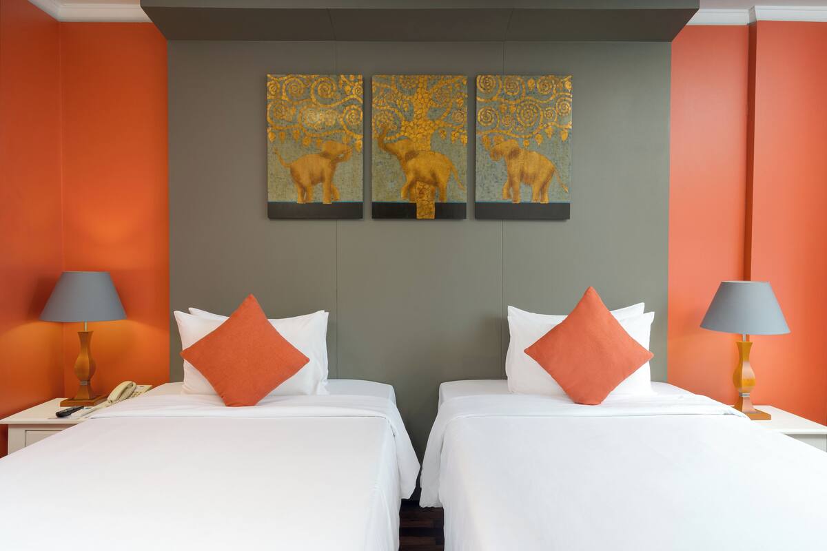 Ein Hotelzimmer mit zwei Betten, weißer Bettwäsche, orangefarbenen Kissen, einem Telefon und einer Lampe auf jeder Seite.