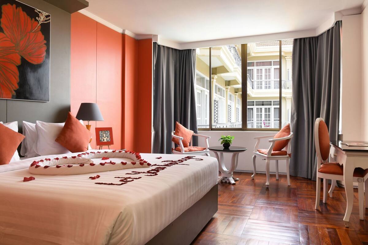 Une chambre d’hôtel avec un grand lit, une petite table ornée d’une plante, deux chaises et une vue sur un immeuble à travers de grandes fenêtres.