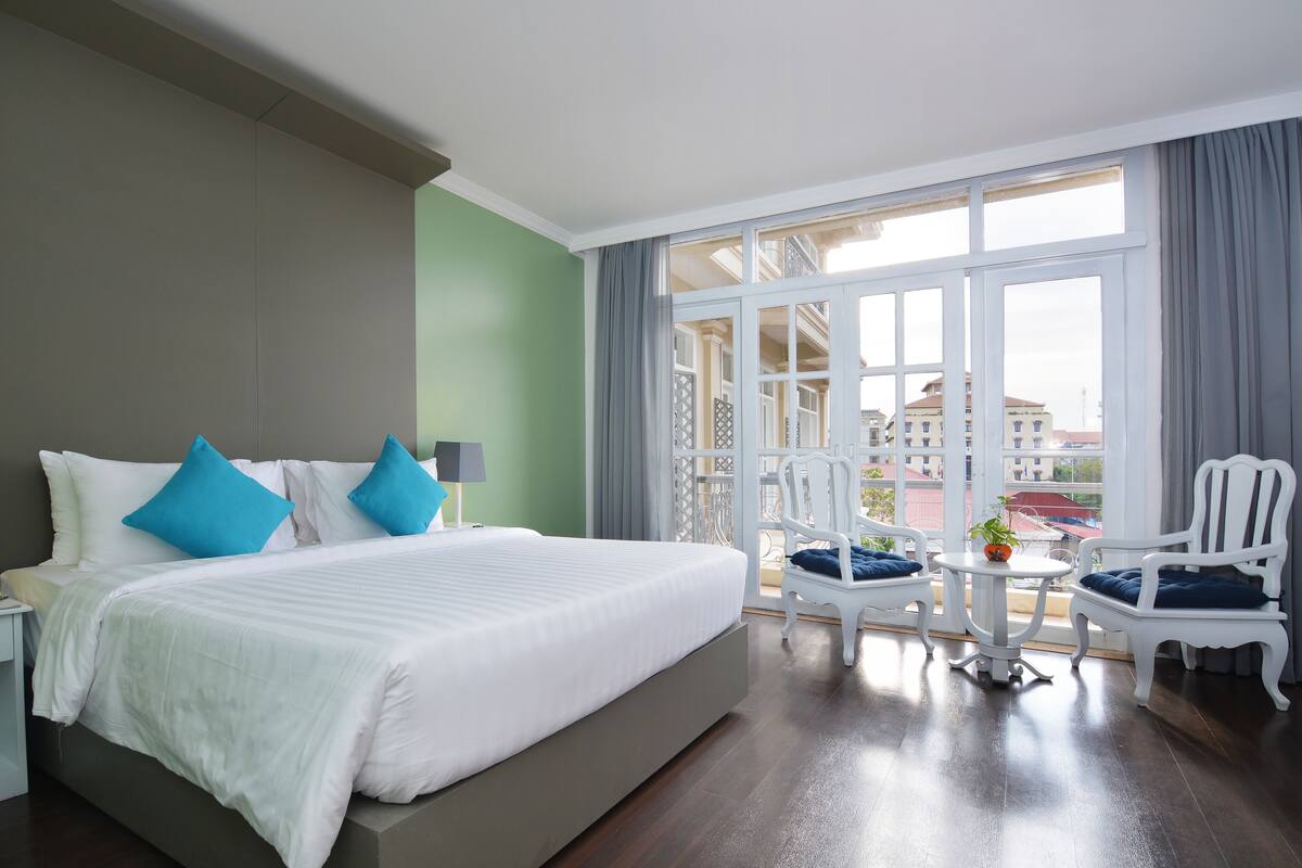 Ein Hotelzimmer mit einem großen Bett, zwei Stühlen, einem kleinen Tisch und Blick auf die Stadt durch große Fenster.