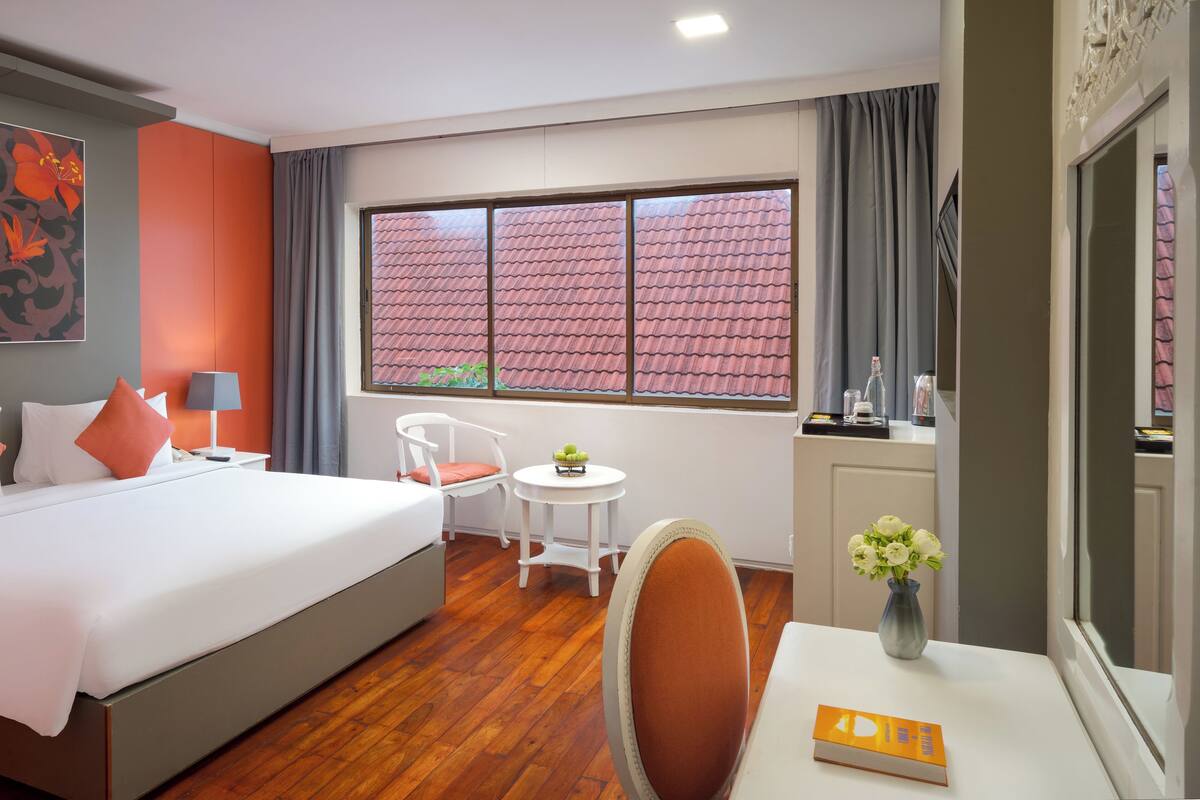 Ein Hotelzimmer mit einem großen Bett, einem kleinen Tisch mit Blumen, einem Sessel, einem Schreibtisch und einem Fenster mit Blick auf ein Ziegeldach.