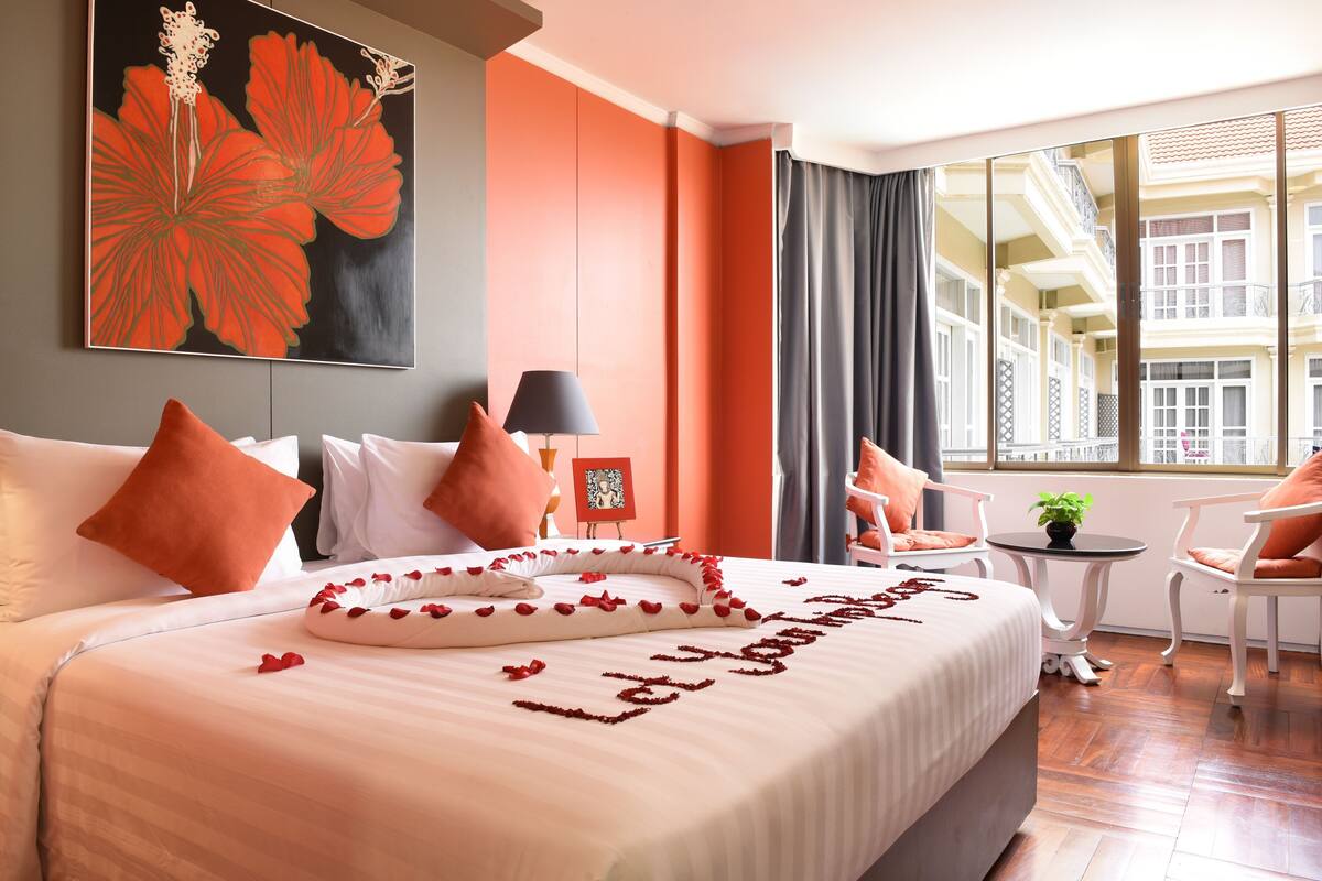 Ein Hotelzimmer mit einem großen Bett, einem Gemälde, einem kleinen Tisch mit Pflanze und Blick auf Gebäude durch das Fenster.