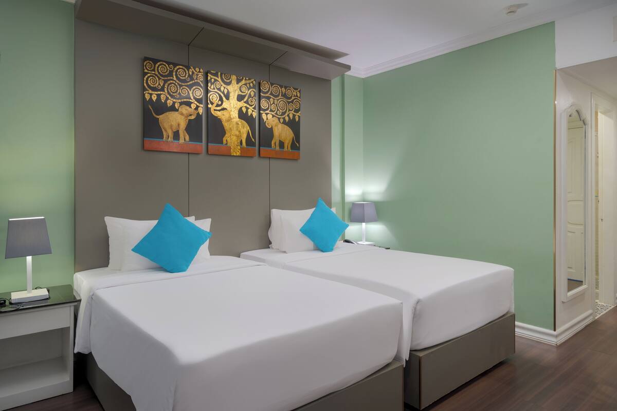Ein Hotelzimmer mit zwei Betten, einem Nachttisch mit Lampe, einem an der Wand hängenden Kunstwerk und einer Tür, die in einen weiteren Raum führt.