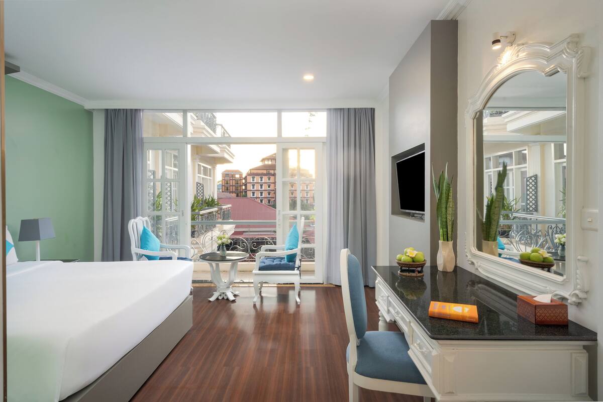 Ein Hotelzimmer mit Bett, Schreibtisch, Stuhl, Fernseher und einem Balkon mit Blick auf Gebäude.