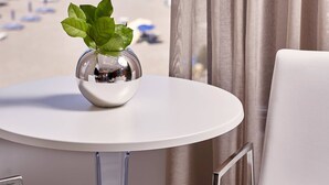Habitación con vista a la playa y al océano. Hay una mesa blanca con un florero de vidrio que contiene una planta.