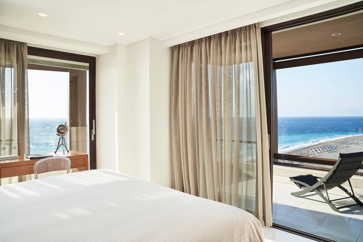Une chambre d’hôtel avec un lit, un bureau doté d’une lampe, une chaise et une vue sur la plage et l’océan, grâce à de grandes fenêtres.