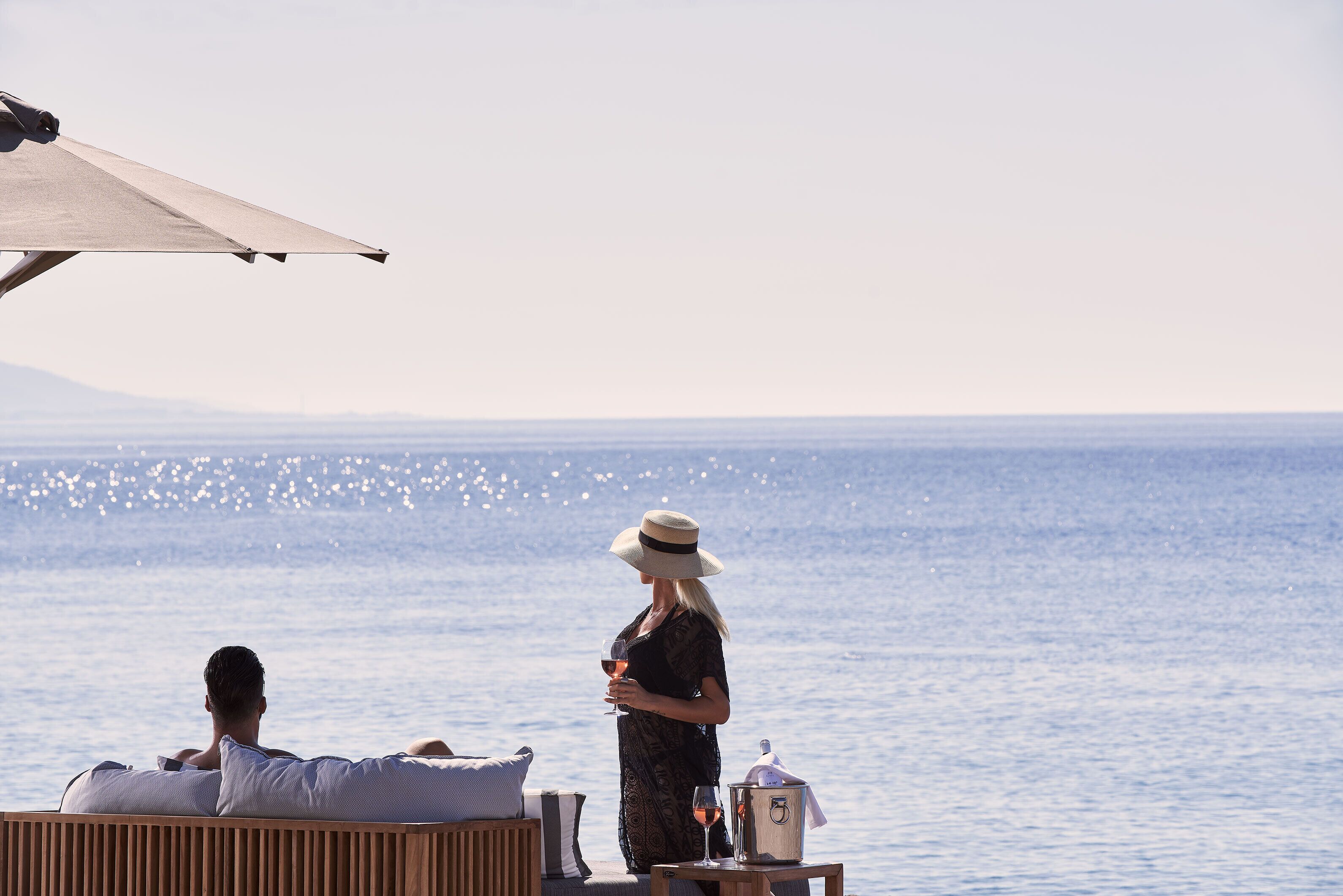 A couple relaxing by a pool with a view of the sea, a lounge chair, and a table with a drink and a bucket.
