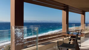 Un balcón con barandilla de vidrio que da al mar, una mesa con sillas y una vista despejada de la playa y montañas a lo lejos.