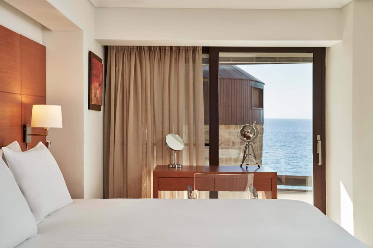 Une chambre d’hôtel avec un lit, un bureau doté d’une lampe, un miroir et une vue sur la mer, accessible par une porte coulissante.