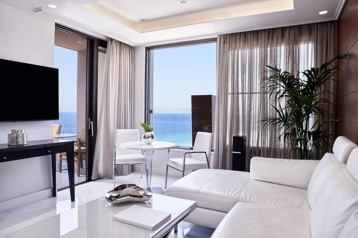 Une chambre d’hôtel moderne avec un canapé en cuir blanc, une table à manger avec des chaises, un téléviseur à écran plat et une grande fenêtre donnant sur la mer.