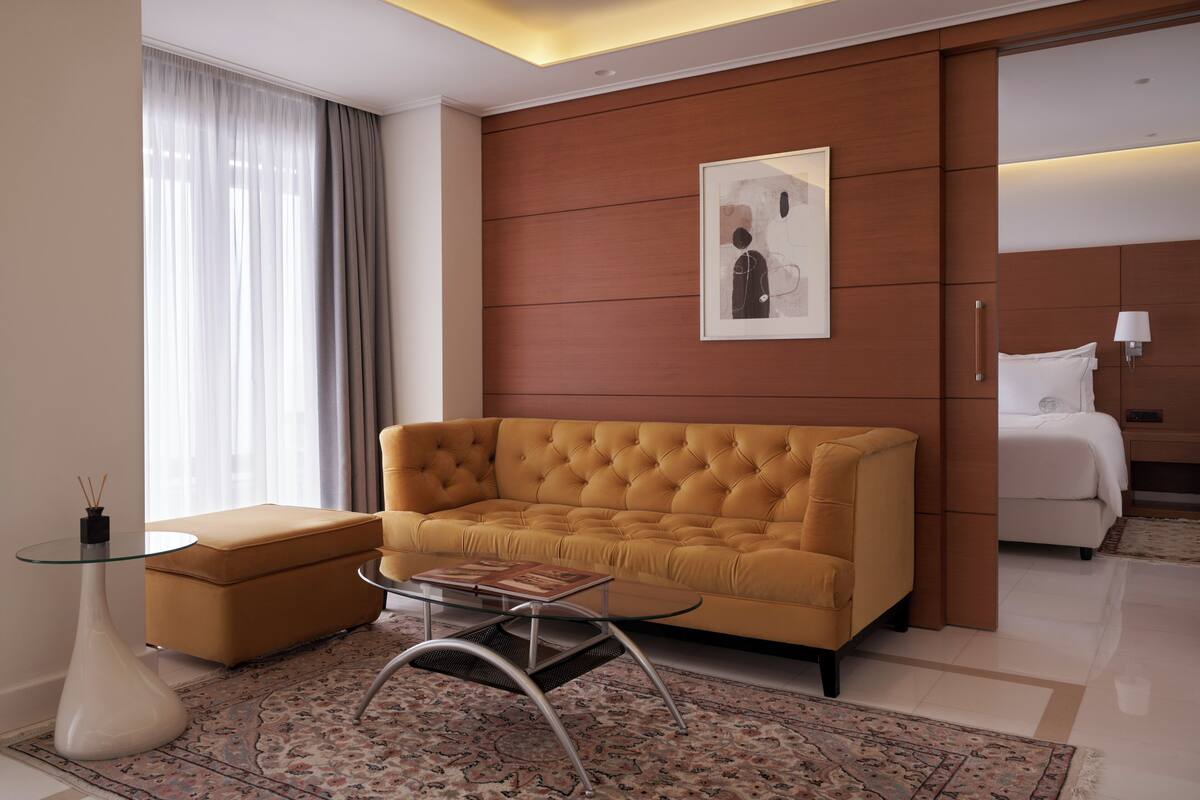 Une chambre d’hôtel moderne avec un canapé capitonné, une table basse en verre, un tapis à motifs, une œuvre d’art encadrée au mur et un lit avec une tête de lit blanche.