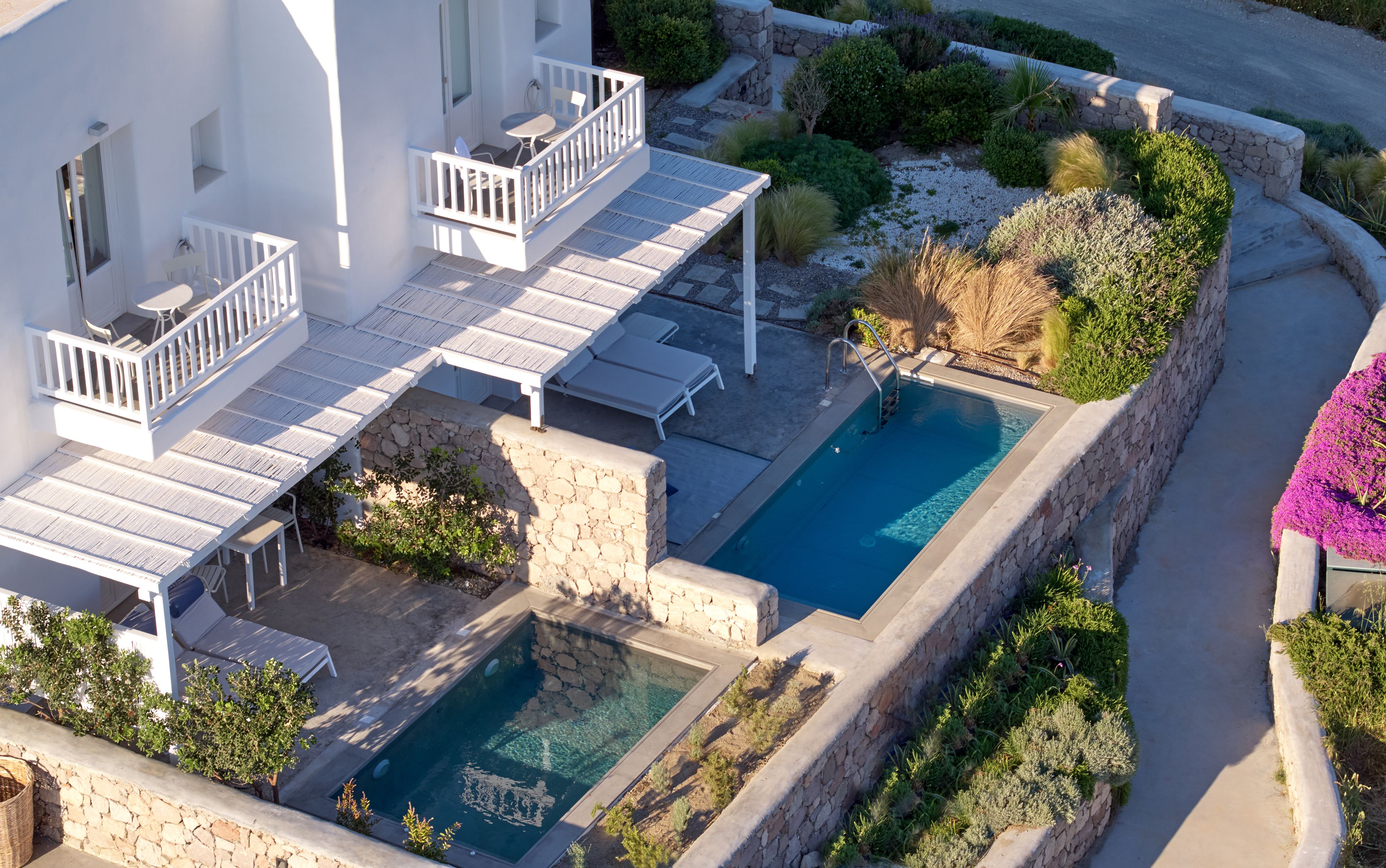 A rooftop pool with a stone wall and lounge chairs, surrounded by greenery and a white building.
