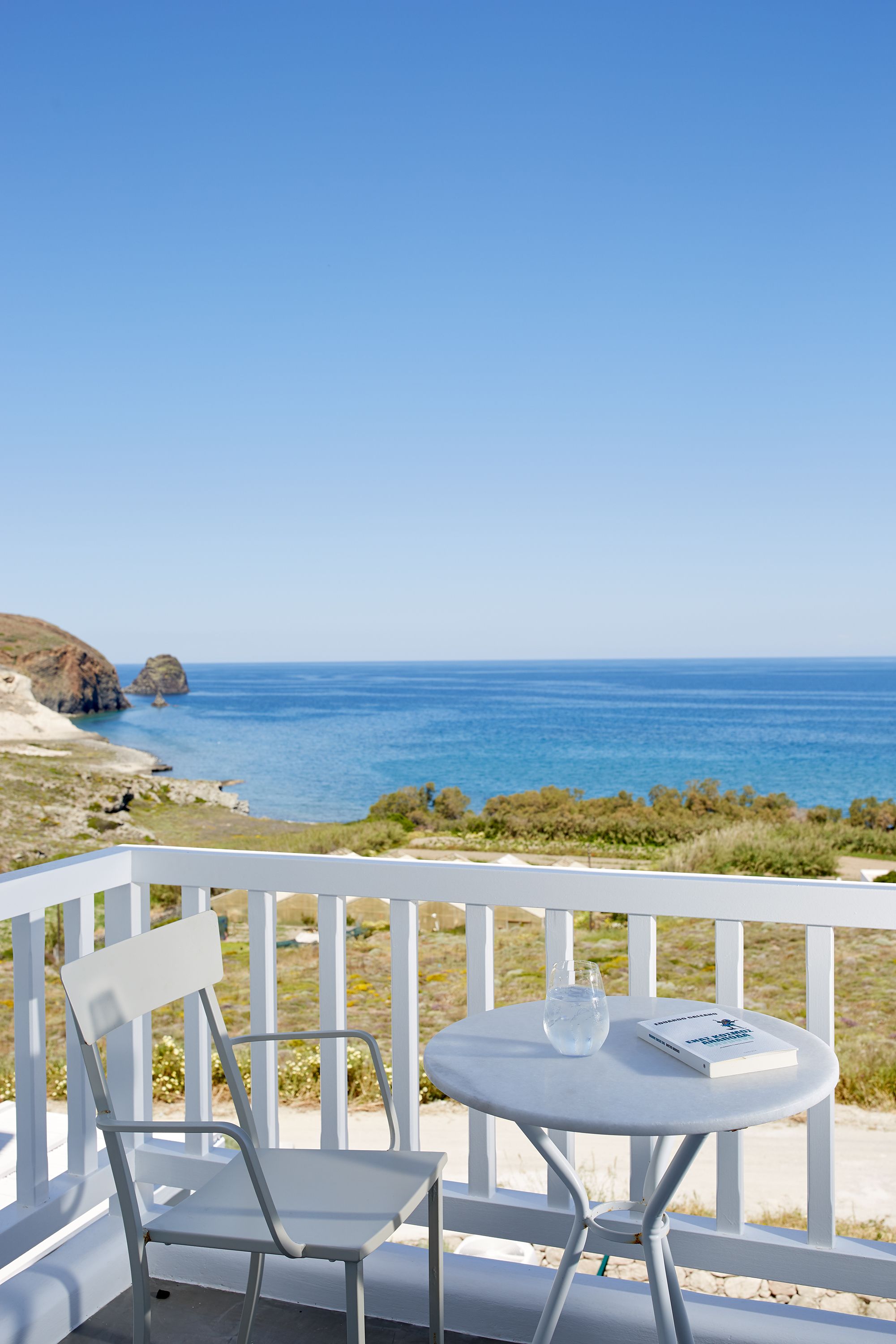 A white metal chair and table on a balcony with a clear view of the ocean and rocky coastline.