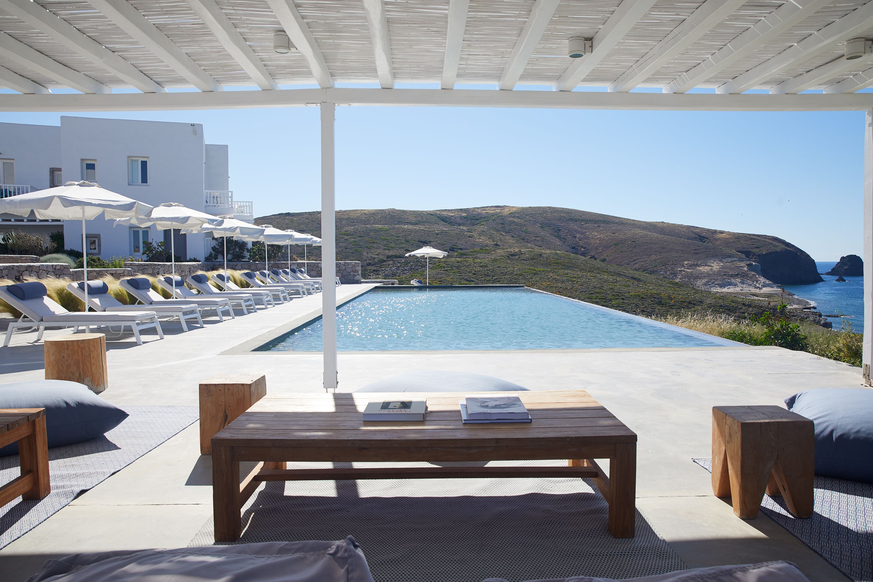 A poolside area with lounge chairs and umbrellas, a wooden table, and a view of the sea and hills.