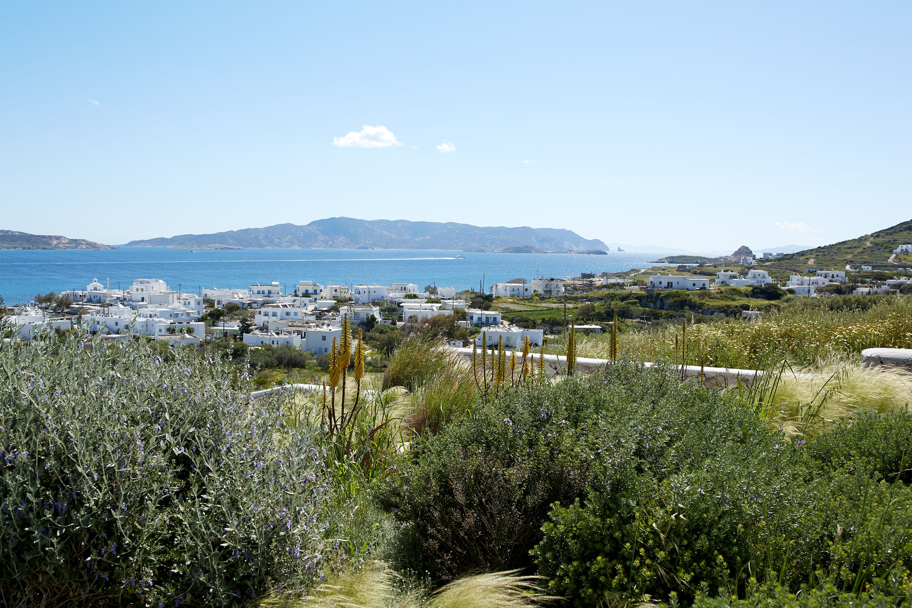 A coastal village with white buildings, a clear blue sea, and distant mountains.