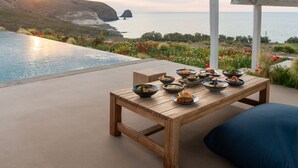 Mesa de madera con cuencos de comida en un patio con vistas a una piscina y al mar.
