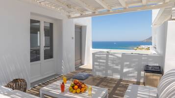 A sunlit patio with a white table set with fruit and drinks, a white sofa, and a view of the sea.