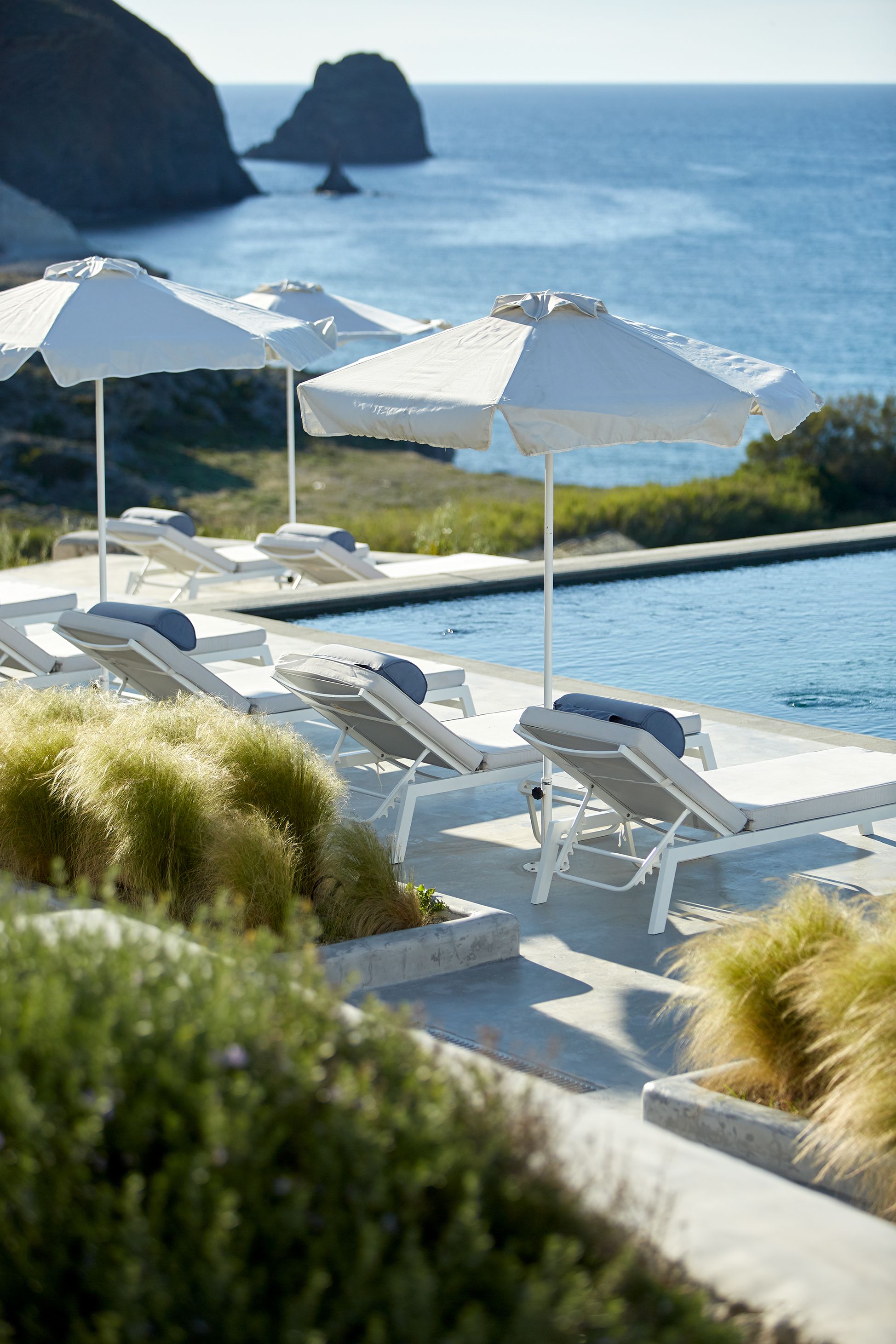 A poolside area with lounge chairs and umbrellas, overlooking a body of water with a rocky outcrop in the distance.