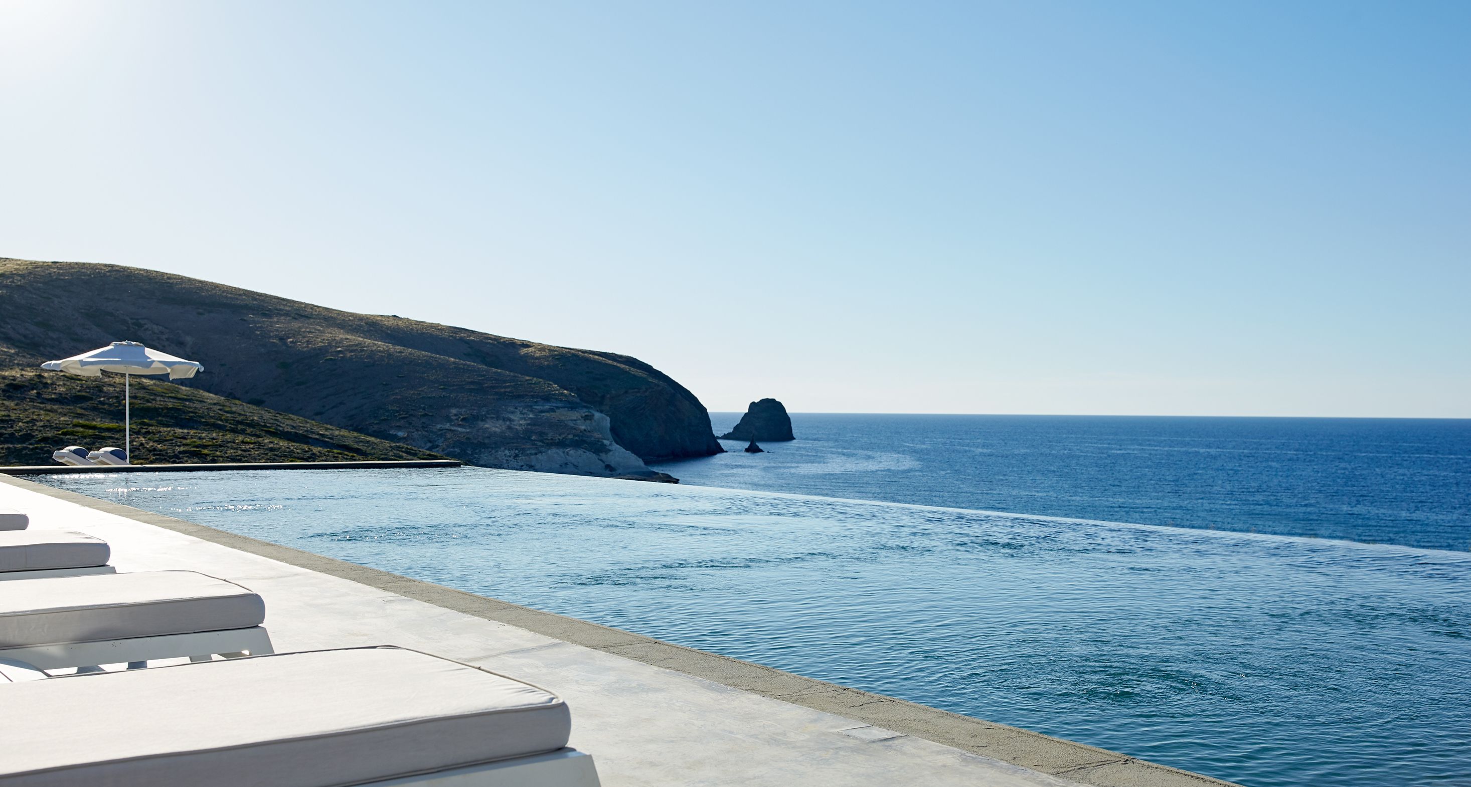 A poolside area with lounge chairs and a white umbrella, overlooking a clear blue sea and a rocky coastline.