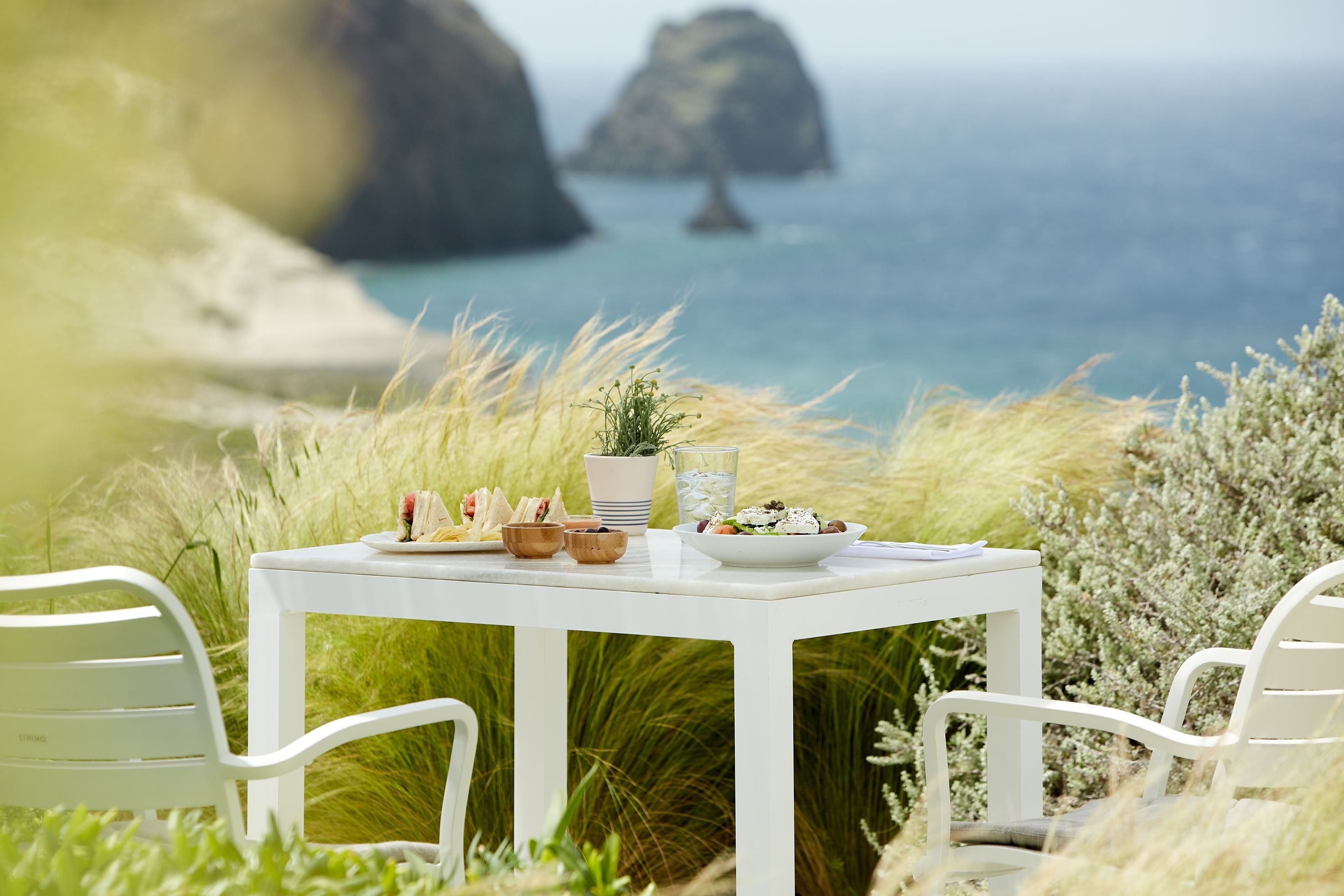 A white table with a meal set and a potted plant, overlooking a coastal landscape with rocky outcrops and the sea.