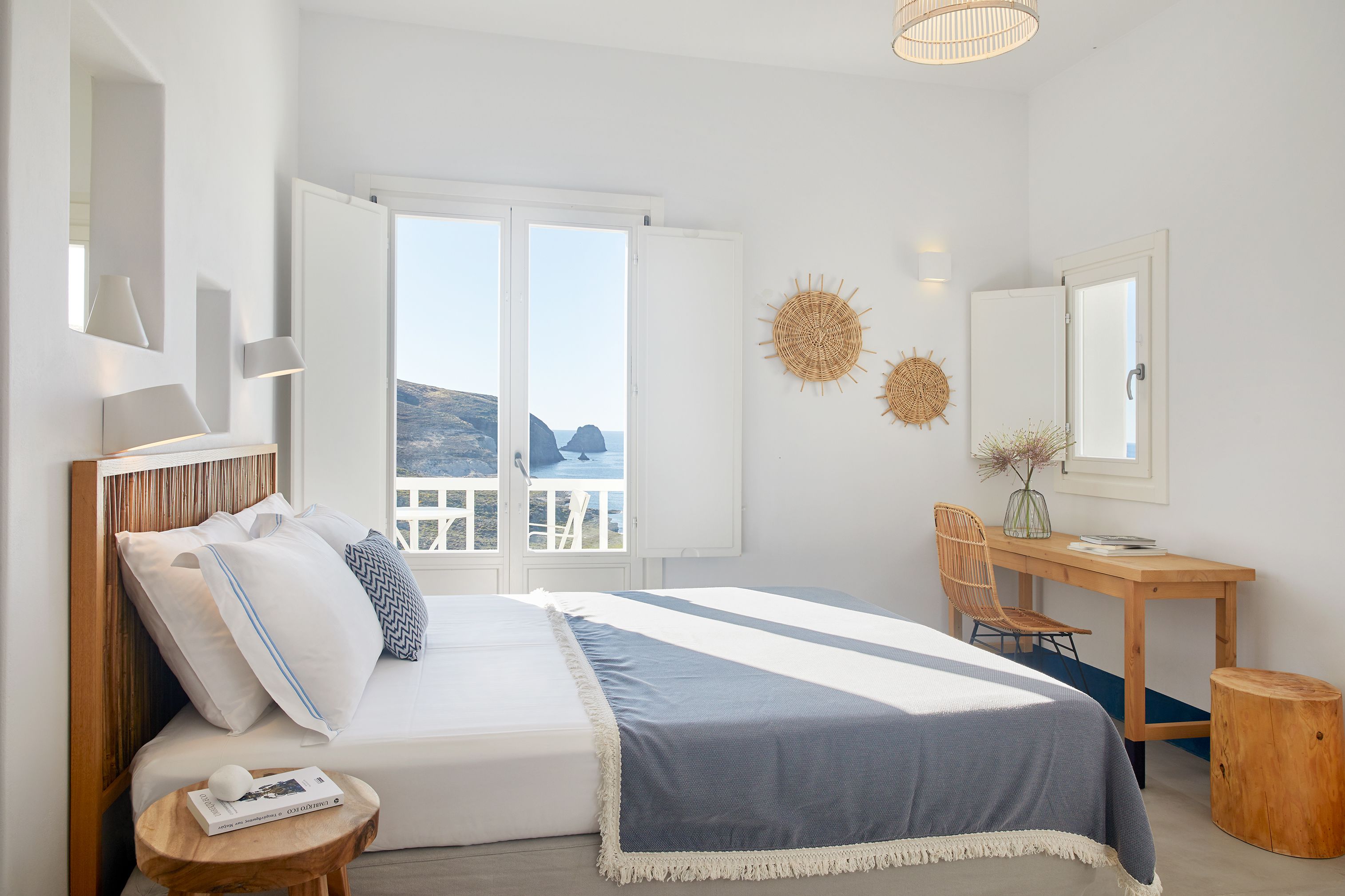 A bedroom with a bed, a desk, and a chair. There is a window with a view of the sea.