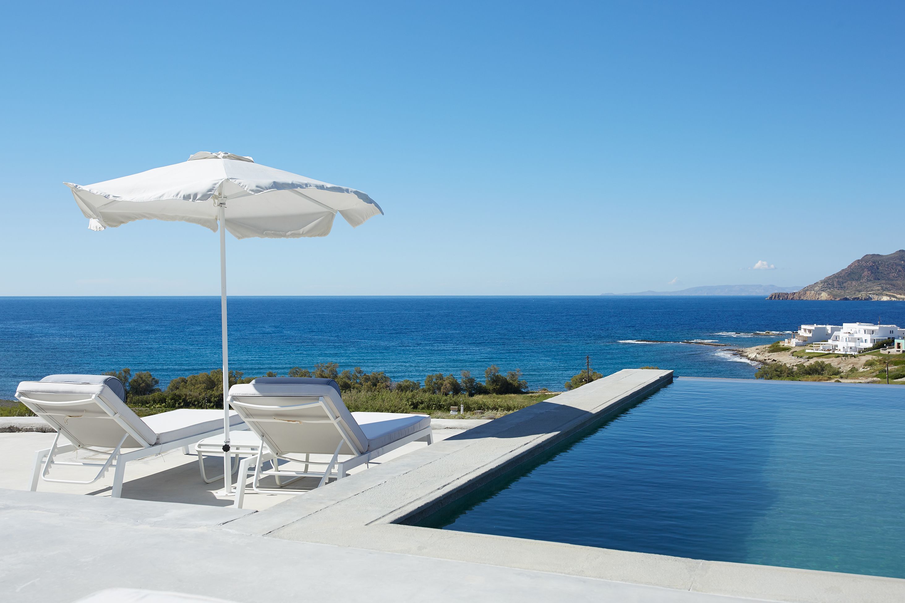 Una piscina infinita con sillones blancos y una sombrilla, con vistas al mar azul cristalino y un paisaje costero.