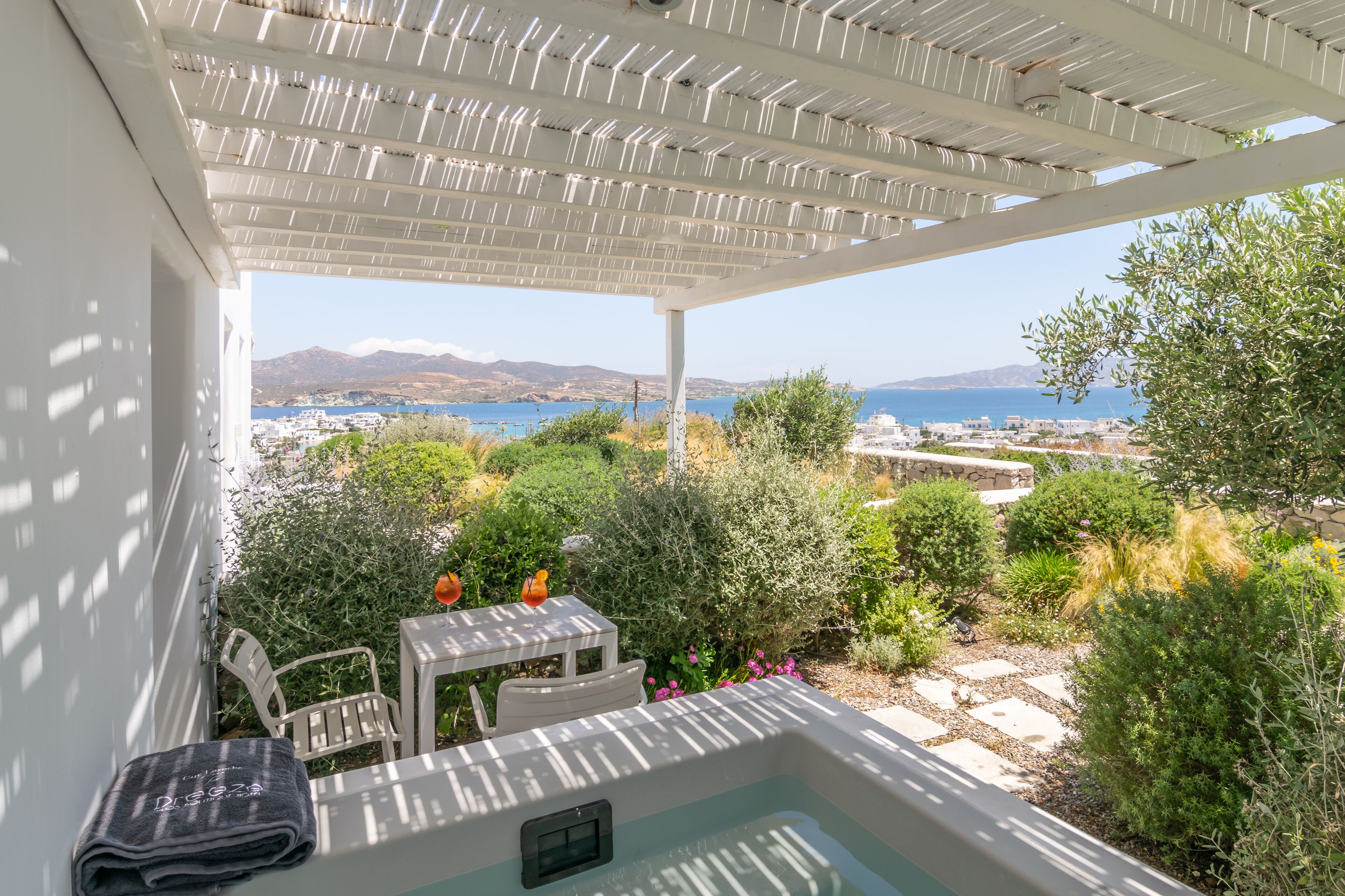A covered outdoor area with a hot tub, white furniture, and a view of the sea and distant buildings.