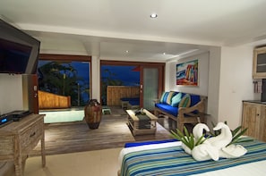 A modern hotel room with a large bed, a sofa, a TV, a wooden coffee table, and a view of a pool and palm trees.