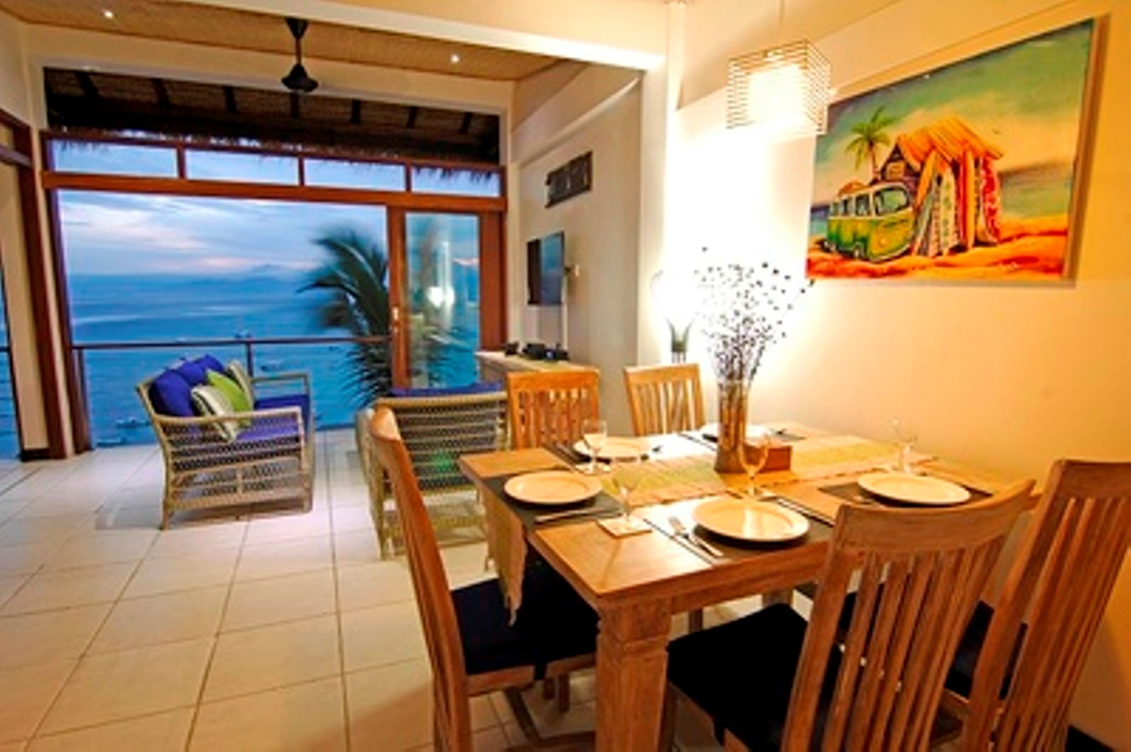 Un coin repas avec vue sur l’océan, un tableau représentant une scène de plage et une table à manger dressée pour quatre personnes.
