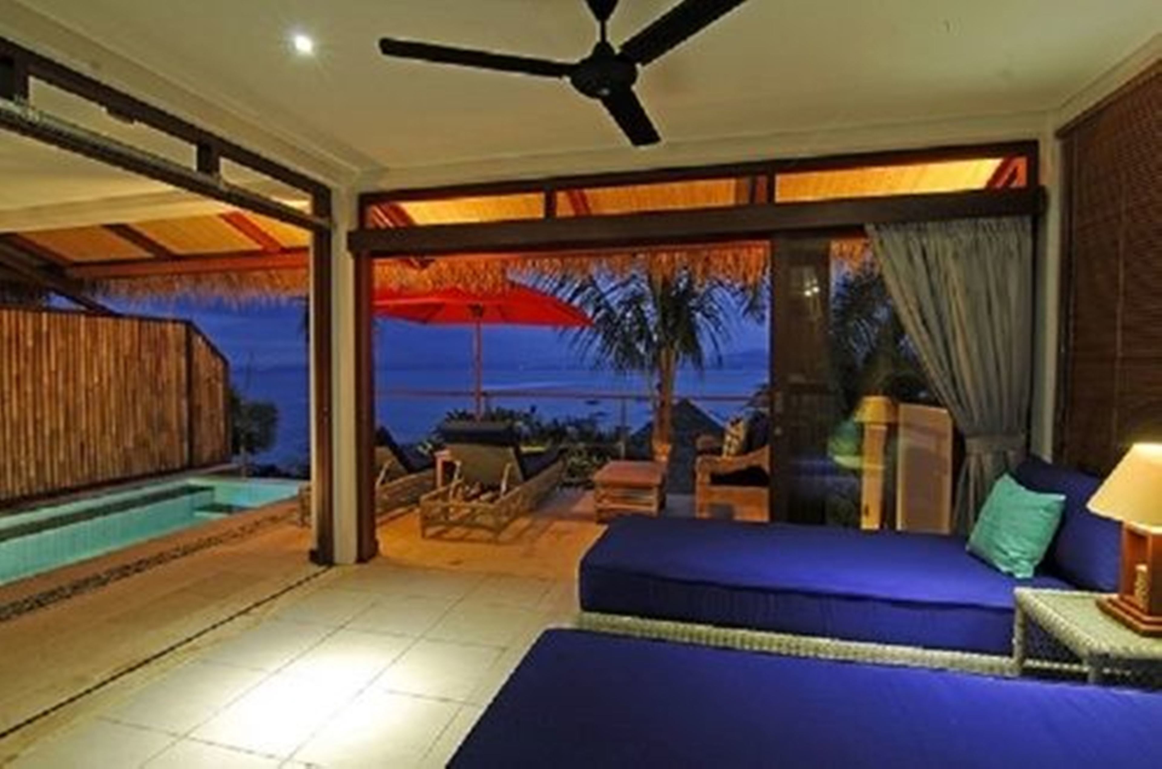 A room with a bed, a ceiling fan, a pool, and a view of the ocean.