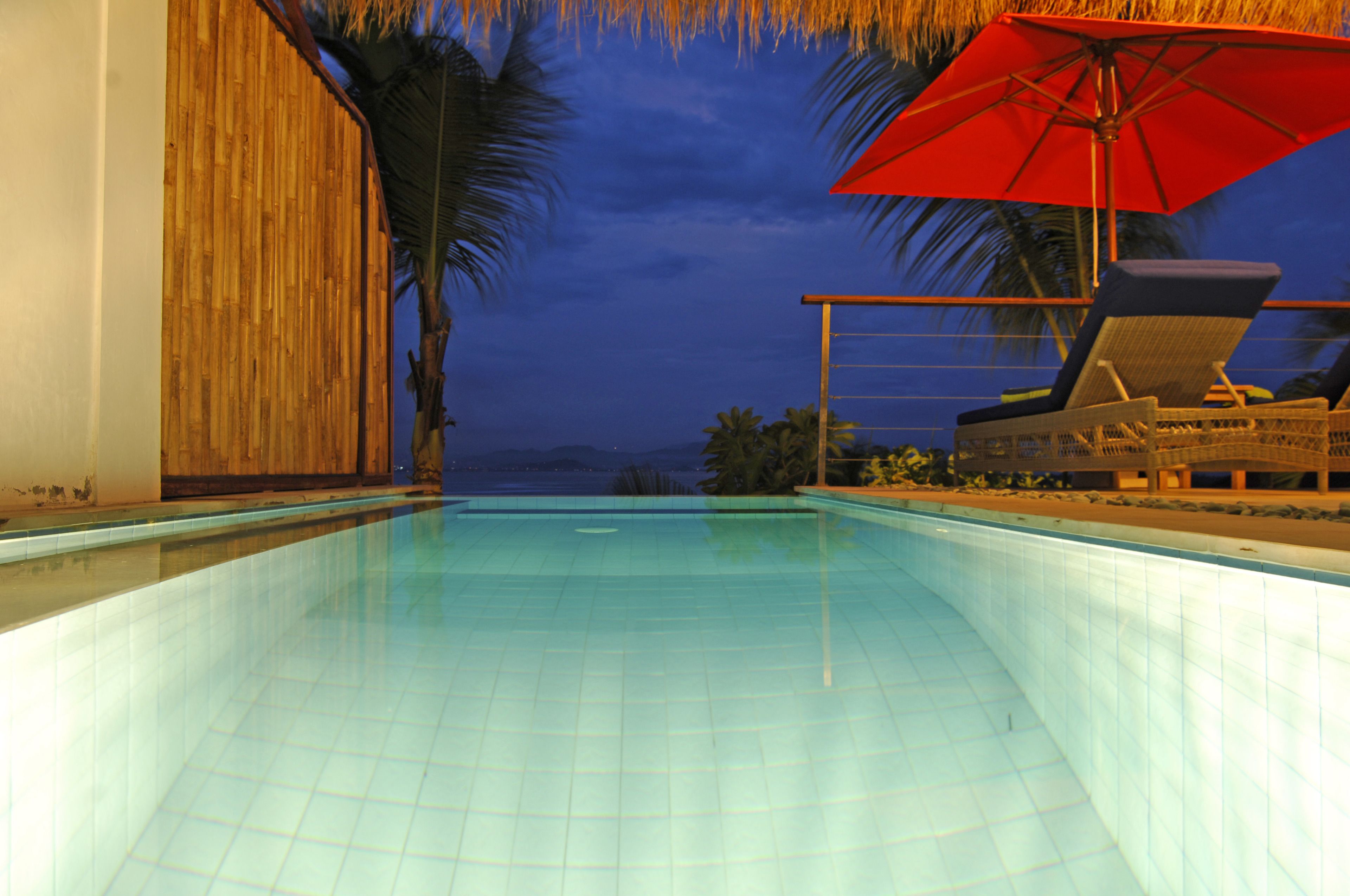 Une piscine avec un parasol rouge, des chaises longues et une vue sur l’océan au crépuscule.