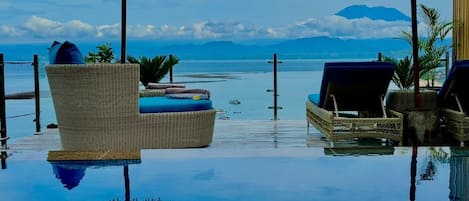 An infinity pool with lounge chairs and umbrellas, surrounded by a clear view of the sea and mountains.